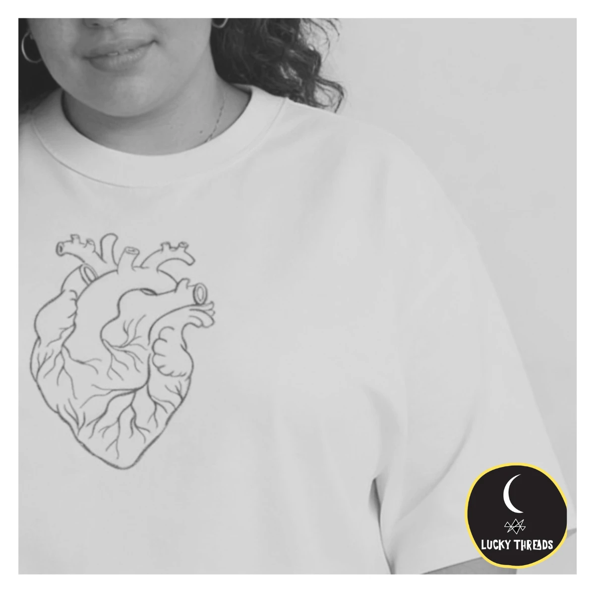 White T‑shirt featuring a black line‑art anatomical heart design printed on the chest, shown on a person from the shoulders to mid‑torso. A small circular “Lucky Threads” logo appears in the bottom corner.