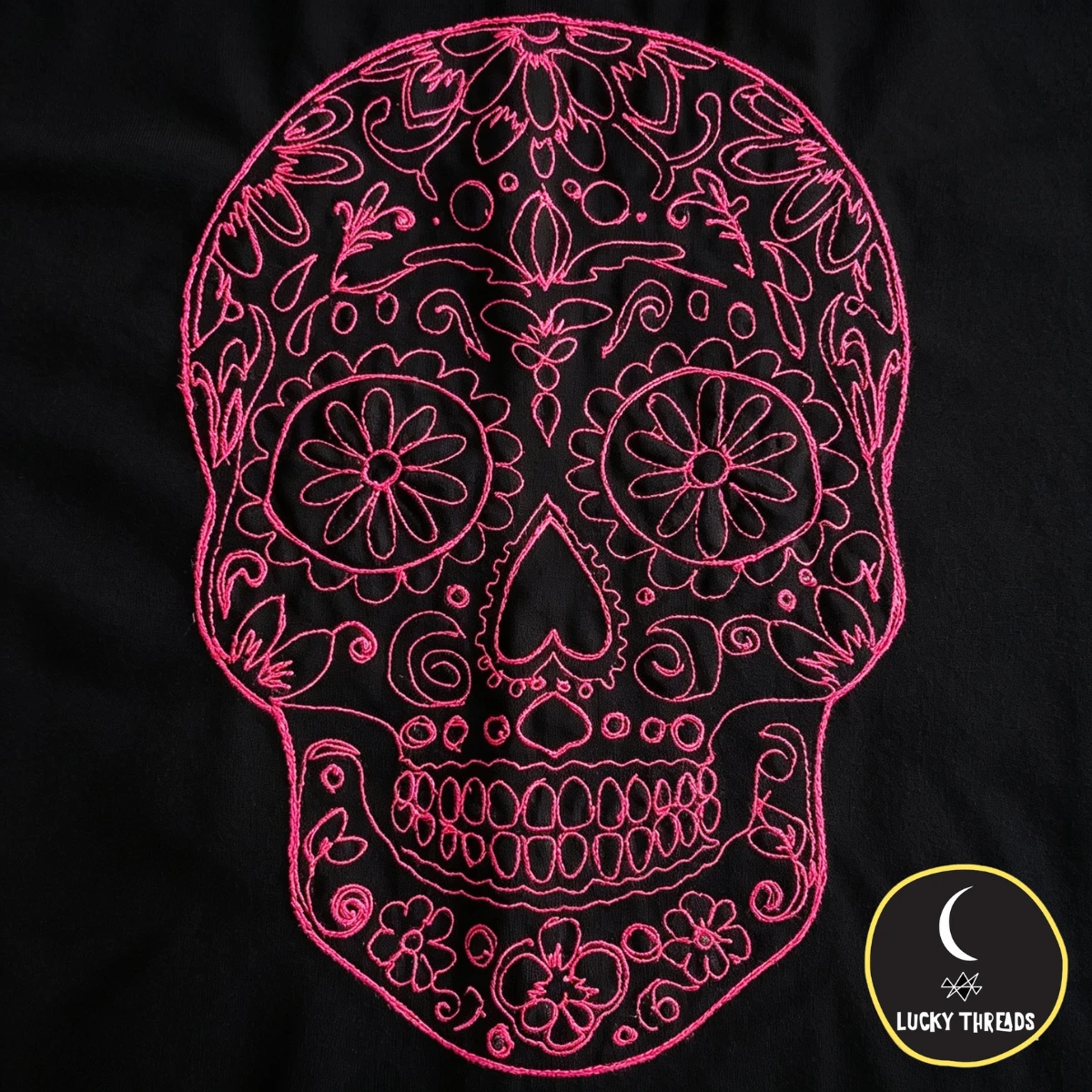 Bright pink embroidered sugar skull design with intricate floral and swirl patterns, stitched onto smooth, deep black fabric; close-up, clean photoshoot style.