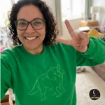 Female customer selfie posing wearing a green sweater with a trceratops stitched design on the front with LuckyThreads logo in bottom right corner.
