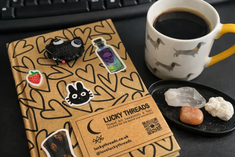 Flat‑lay of a creative workspace featuring a Lucky Threads notebook decorated with stickers, a branded business card, a dachshund‑patterned coffee mug, and a small dish of crystals beside a keyboard.