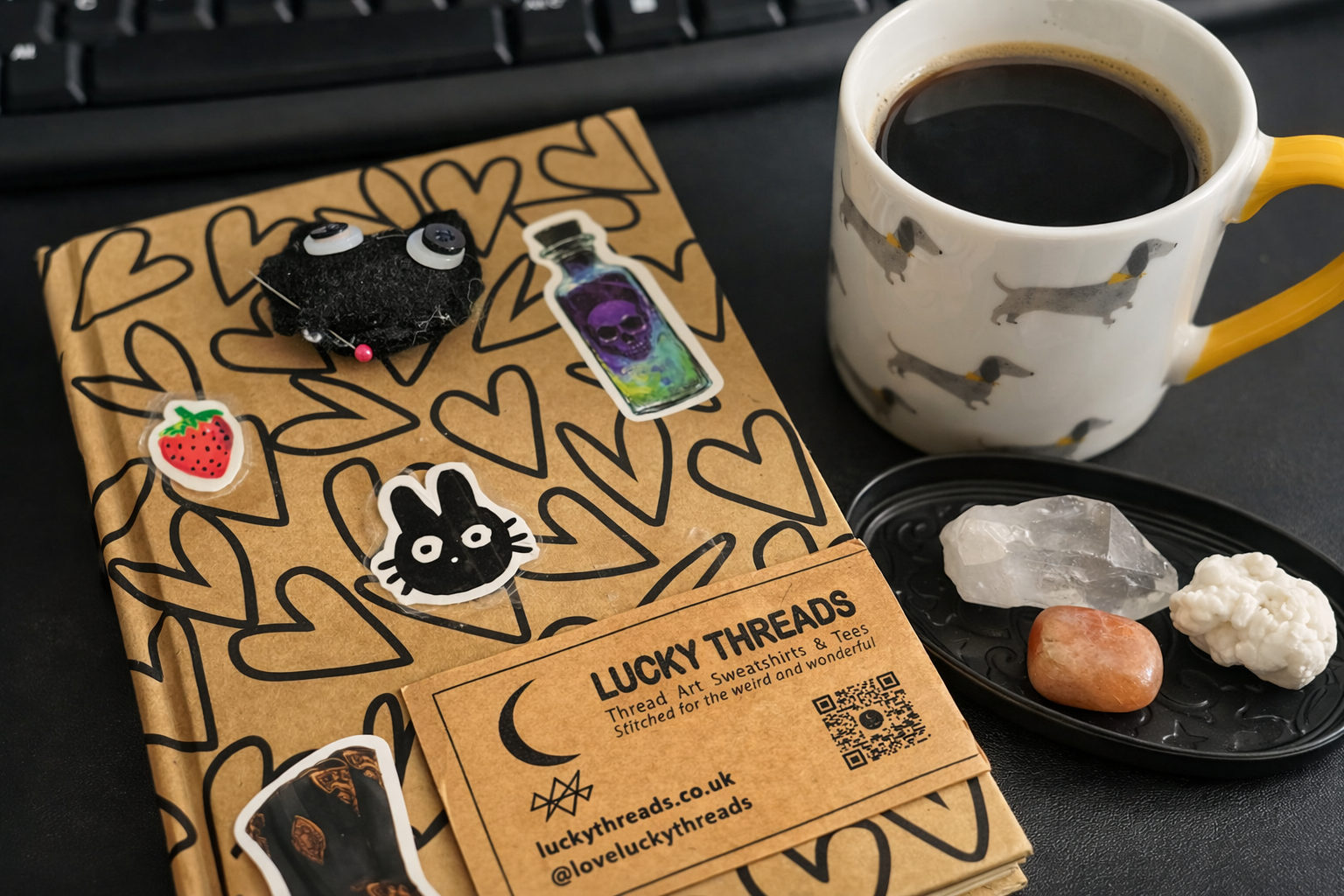 Flat‑lay of a creative workspace featuring a Lucky Threads notebook decorated with stickers, a branded business card, a dachshund‑patterned coffee mug, and a small dish of crystals beside a keyboard.